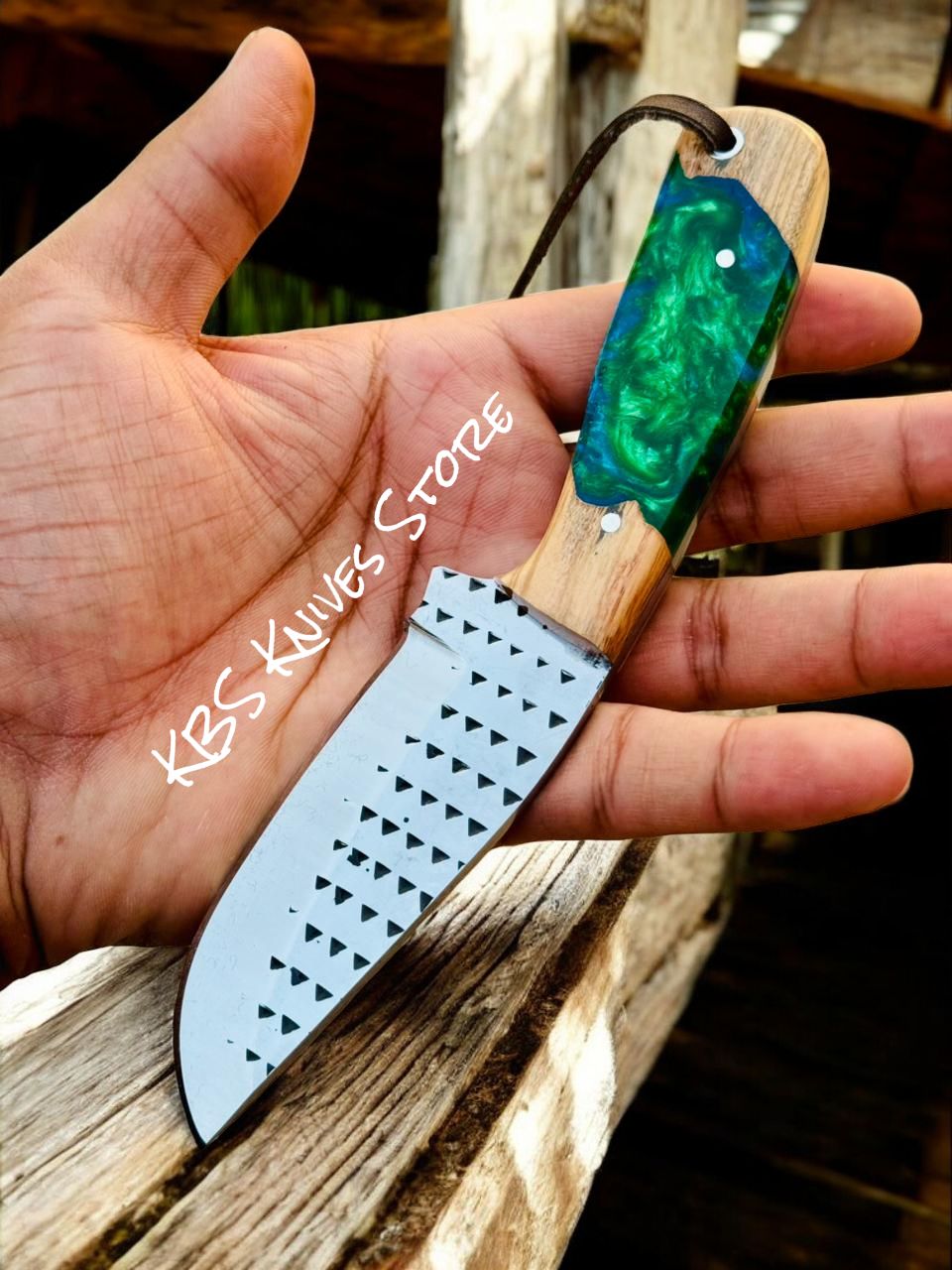 Hand holding a small knife with a green handle and decorative blade on a wooden surface.