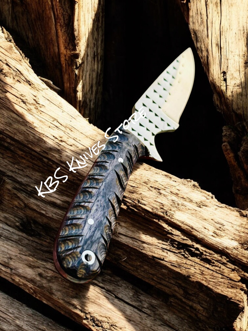 Knife with a textured handle on wooden logs, branded 'KBS Knives & Tools'.