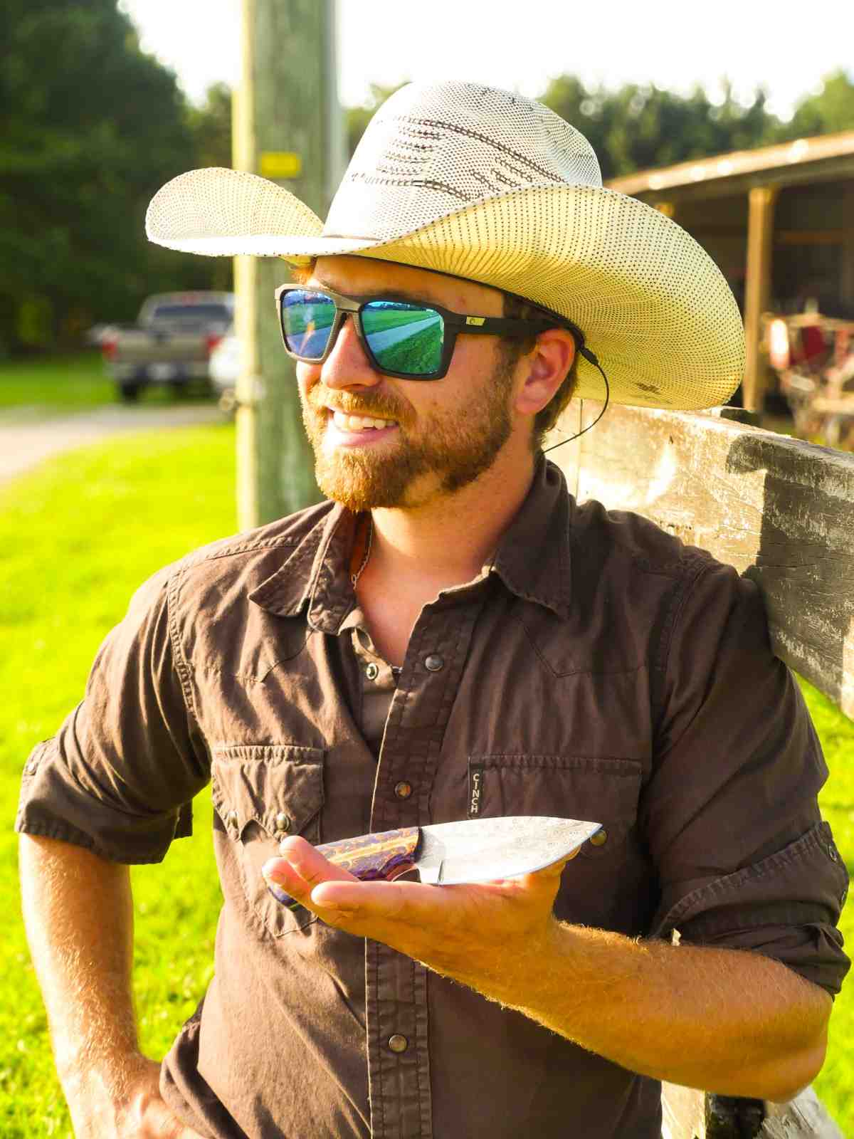 Man wearing a straw hat and sunglasses, holding a metal object in a grassy outdoor setting.