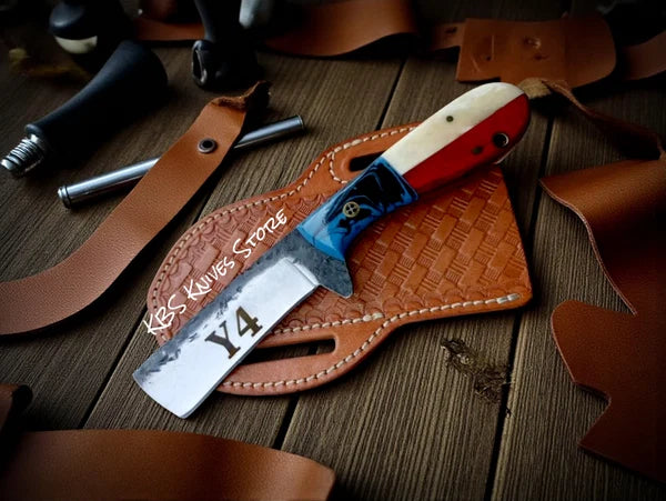 A custom fixed-blade knife with a silver blade, red and cream handle, and blue guard, resting on a brown leather sheath over a wooden surface. The blade is engraved with 'Y4' and the handle features a skull graphic.