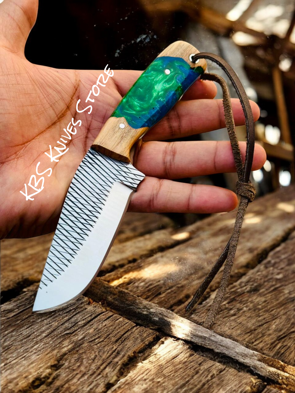 Hand holding a knife with a decorative handle on a wooden surface, branded 'KBS Knives Store'.