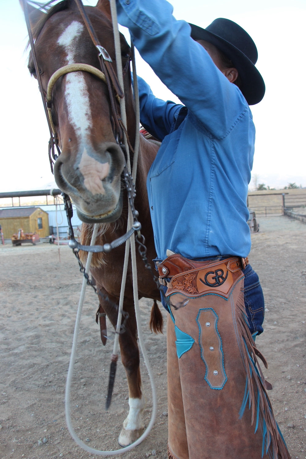 Man in blue shirt and cowboy hat adjusting a horse's bridle in an outdoor setting.