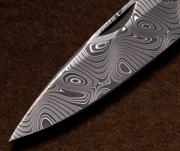 A close-up of a Damascus steel knife blade with a distinctive wavy, swirling pattern of black and silver layers, resting on a dark brown textured surface.