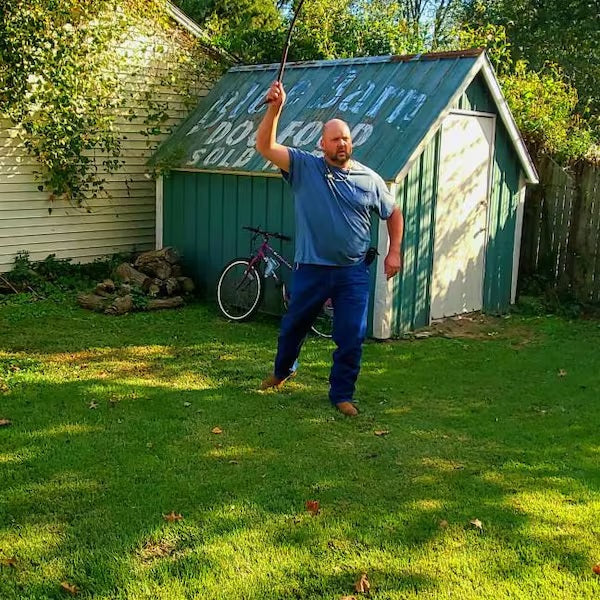 Man in a blue shirt and pants standing in a grassy area with a green shed and bicycle in the background.
