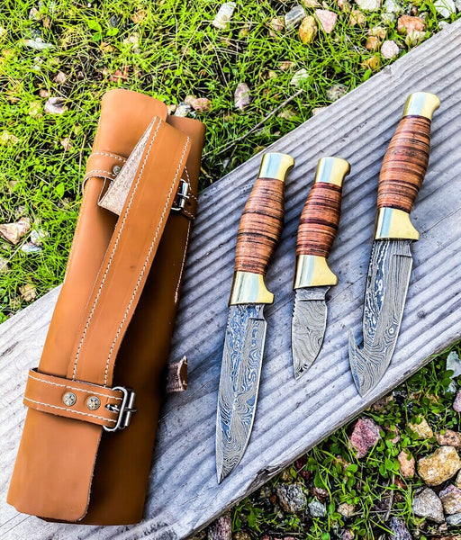 Image showing the Wilderness Trio Hunting Knives Set, featuring Guthook, Hunting, and Skinning knives with Damascus steel blades, stacked leather handles, brass guards, and presented with a leather roll. Essential tools for outdoor enthusiasts.
