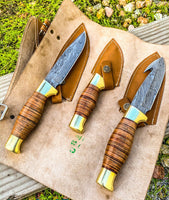 Image showing the Wilderness Trio Hunting Knives Set, featuring Guthook, Hunting, and Skinning knives with Damascus steel blades, stacked leather handles, brass guards, and presented with a leather roll. Essential tools for outdoor enthusiasts.