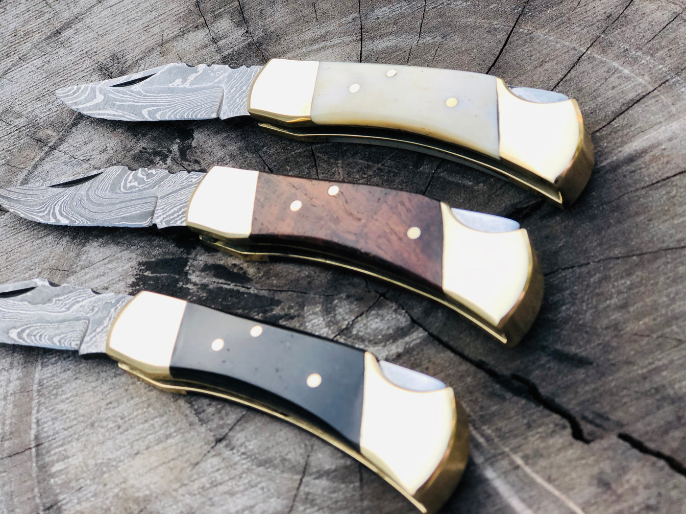 A trio of exquisite folding pocket knives with Damascus steel blades, featuring handles made from buffalo horn, bone, and rosewood, adorned with brass bolsters. Each knife measures 5 inches overall and comes with a stylish leather case for secure storage and portability.